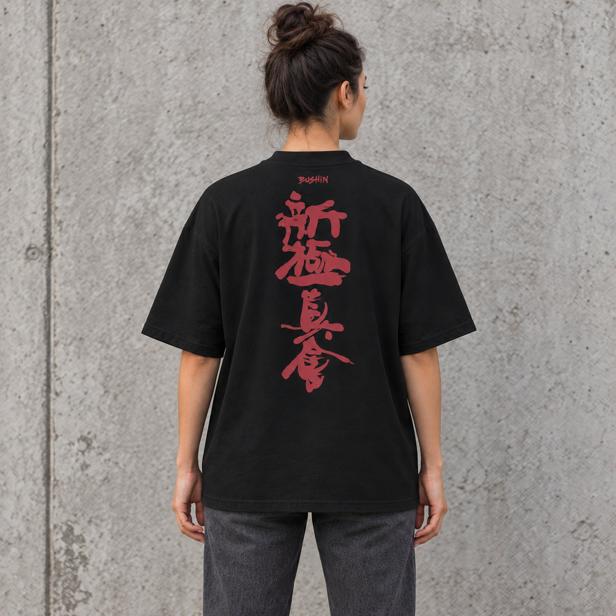 Back view of a female model wearing a black BUSHIN oversized T-shirt with crimson Shinkyokushin kanji against a grey concrete wall.