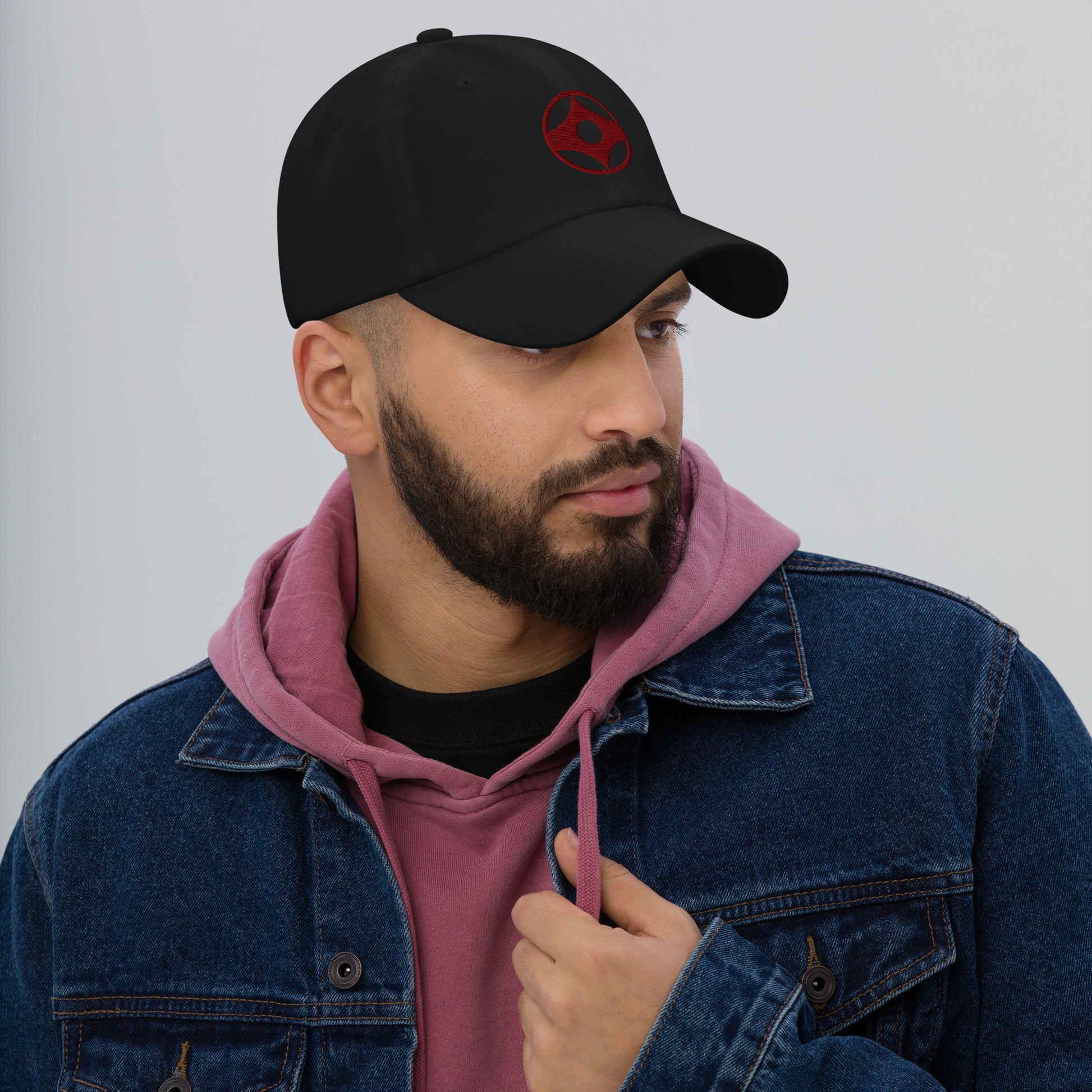 Man wearing a black cap with text, pink hoodie, and blue denim jacket on a gray background