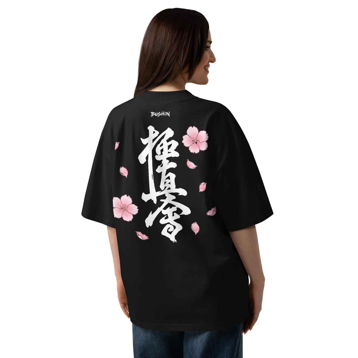 A woman shown from the back wearing an oversized black T-shirt with large white Japanese calligraphy and pink cherry blossom graphics on the back, photographed against a plain white studio background.