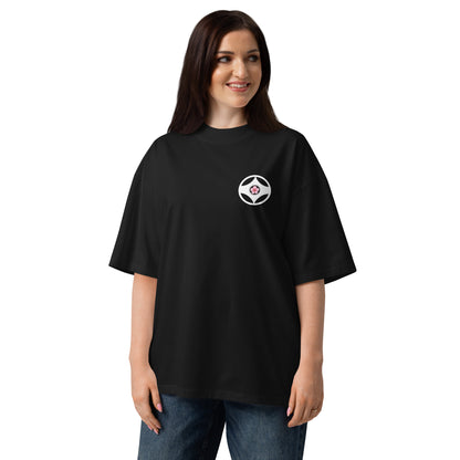 A woman modeling an oversized black t-shirt with a small circular white and red logo on the left chest, set against a plain white background.