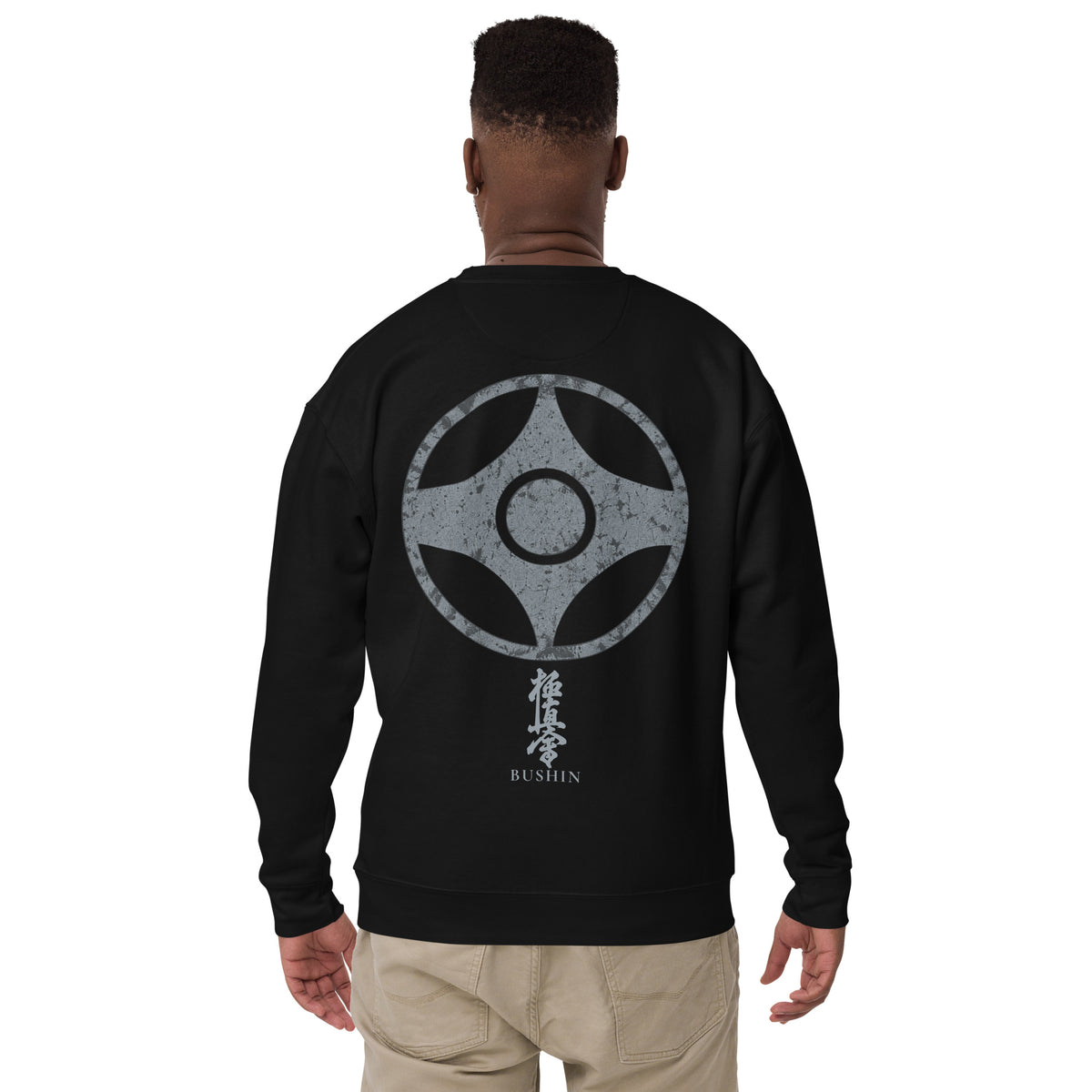 A man viewed from the back wearing a black crewneck sweatshirt with a large grey kyokushin kanku graphic print on the back, paired with khaki pants, against a plain white background.